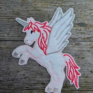 Peut inclure: Applique en feutre blanc en forme de licorne avec des broderies roses et argentées. La licorne a une corne argentée, une crinière et une queue roses, et des ailes argentées.