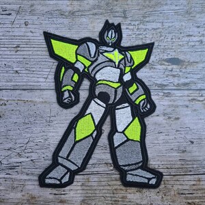 Könnte beinhalten: Ein grauer und weißer Roboter mit gelben Akzenten und einem Stern auf der Brust. Der Roboter steht mit seinen Armen an den Seiten und seinen Beinen auseinander. Der Roboter befindet sich auf einem hölzernen Hintergrund.