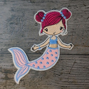May include: Embroidered patch of a cartoon mermaid with pink hair in two buns, blue details, and a pink and blue tail. The mermaid wears a blue top and has a smiling face. The patch is edged in white.