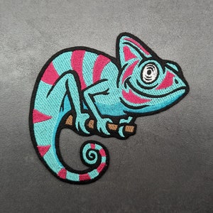 May include: A blue and pink embroidered chameleon patch with a whimsical expression. The chameleon is curled up with its tail wrapped around its body.