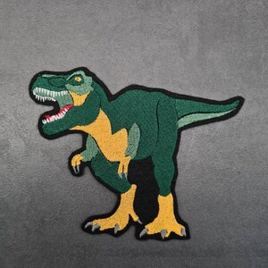 May include: Green and yellow embroidered dinosaur patch with a black outline. The dinosaur is standing on its hind legs with its mouth open and teeth showing.