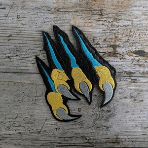 Könnte beinhalten: Gestickte Krallen-Patches mit schwarzer Basis und blauen, gelben und grauen Akzenten. Die Patches sind auf einer verwitterten Holzoberfläche angeordnet. Diese Patches eignen sich für Jacken, Taschen oder andere Stoffartikel.