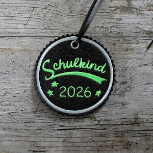 Może przedstawiać: Czarna, okrągła, haftowana naszywka z napisem "Schulkind 2026" w zielonym skrypcie i gwiazdkami. Naszywka ma białą obwódkę i czarną wstążkę do zawieszenia. Tło to zwietrzała drewniana powierzchnia.