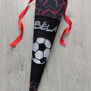 Könnte beinhalten: Ein schwarzer und roter Schultüte mit einem Fußball-Design und dem Namen "Béla" in silbernen Lettern.