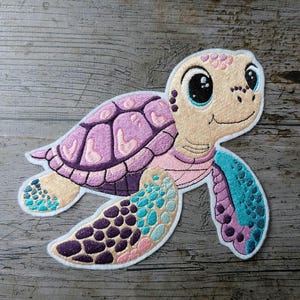 Pode incluir: Remendo bordado de uma tartaruga marinha sorridente. A tartaruga tem um rosto castanho claro, uma carapaça rosa e roxa e barbatanas azuis e bege. O remendo tem uma borda branca.