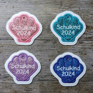 May include: Four seashell-shaped patches with the text "Schulkind 2024". Each patch has a white border and a glittery interior in pink, teal, purple, and blue. The patches are likely for a school-related event.