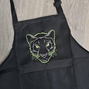 May include: Black apron with a green embroidered panther head design. The apron has a large pocket on the front.