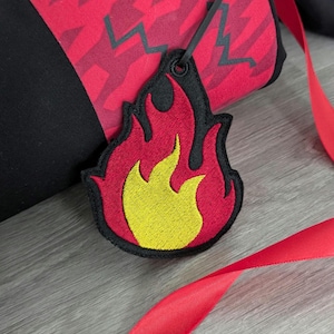 May include: A red and black fabric bag with a black strap and a yellow and red embroidered flame charm.