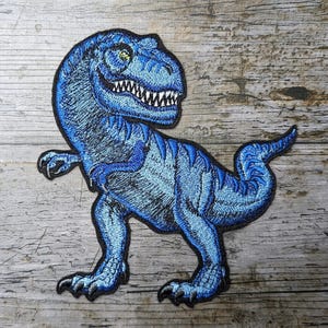 May include: Embroidered patch of a blue Tyrannosaurus Rex dinosaur. The patch has a detailed design with varying shades of blue, white teeth, and green eyes. The dinosaur is depicted in a dynamic pose, with its tail curved and one arm raised.