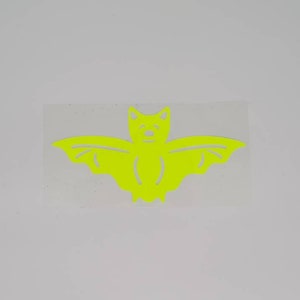 Puede incluir: Una silueta de murciélago amarillo brillante recortada en papel. El murciélago tiene un diseño simple con alas grandes y una cabeza pequeña con dos ojos.