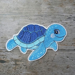 Peut inclure: Écusson brodé d'une tortue de mer bleue souriante avec une carapace et des nageoires bleu clair. La tortue a un contour blanc et un visage bleu clair avec une joue rose. L'écusson est sur une surface en bois.