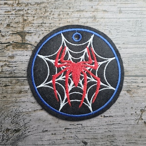 Op de afbeelding: Geborduurde ronde patch met een rode spin en wit webdesign op een zwarte achtergrond. De patch heeft een blauwe rand en een klein gaatje aan de bovenkant. Geschikt voor kleding of accessoires.
