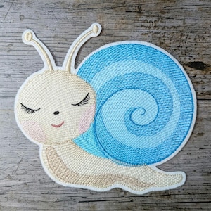 Könnte beinhalten: Eine gestickte Schnecke in Blau und Weiß mit geschlossenen Augen und einem lächelnden Mund. Die Schnecke befindet sich auf einem hölzernen Hintergrund.