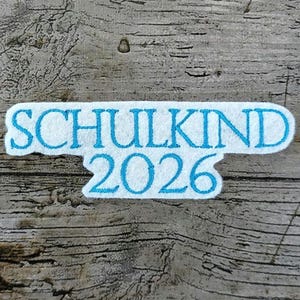 Könnte beinhalten: Ein weißes Filz-Patch mit den Worten "SCHULKIND 2026" in hellblau gestickt. Der Aufnäher hat eine wellenförmige, unregelmäßige Form und ist vor einem strukturierten Holzgrund platziert.