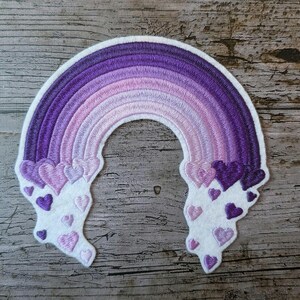 Puede incluir: Parche bordado con un diseño de arcoíris púrpura y borde blanco. El arcoíris transiciona de púrpura oscuro a claro, con acentos en forma de corazón en los extremos. Para ropa o accesorios.