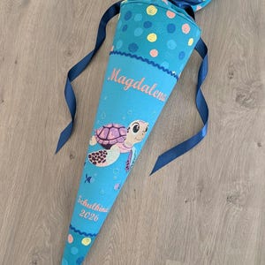 Könnte beinhalten: Eine türkisfarbene Schultüte mit einer Schildkrötenillustration und dem Namen "Magdalena". Die Tüte ist mit blauen, gelben und rosa Punkten und einem blauen Band verziert. Der Text "Schulkind 2026" ist ebenfalls sichtbar.