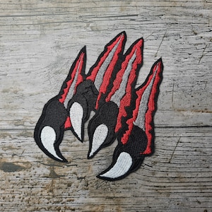 May include: Red and gray embroidered patch of a set of claws. The claws are black with white tips and are arranged in a semi-circle.