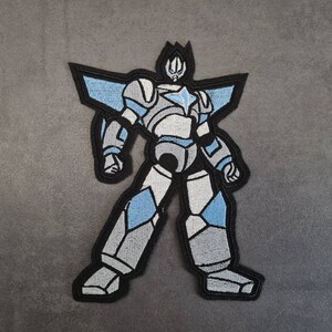 Op de afbeelding: Een geborduurde patch met een robot in zwart en wit, met een blauwe ster op zijn borst. De robot heeft een puntige kop en staat met gespreide benen.