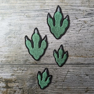 May include: Four embroidered patches shaped like dinosaur footprints. The patches are green with black outlines and white accents. They are arranged on a weathered, grey wooden surface.