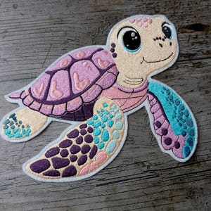 May include: Embroidered sea turtle patch with a smiling face, a pink and purple shell, and blue and purple flippers. The patch is on a wooden surface. The turtle's design includes various shades of pink, purple, blue, and beige.