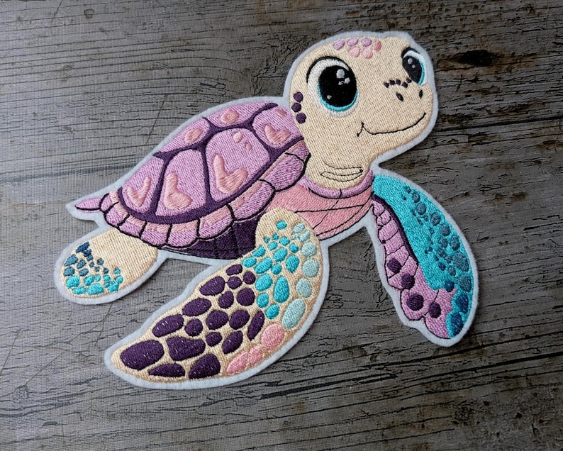 May include: Embroidered sea turtle patch with a smiling face, a pink and purple shell, and blue and purple flippers. The patch is on a wooden surface. The turtle's design includes various shades of pink, purple, blue, and beige.