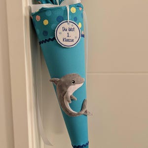 Könnte beinhalten: Eine türkisfarbene Schultüte mit einem Delfin-Applikation und einem gepunkteten Oberteil. Der Kegel hat ein Etikett mit der Aufschrift "Du bist 1. Klasse". Weißes Band ist um die Oberseite gebunden.