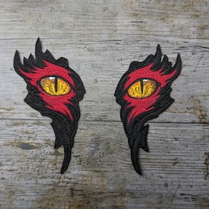 May include: Two embroidered patches depicting a pair of red and black eyes with yellow pupils. The eyes are set against a woodgrain background.