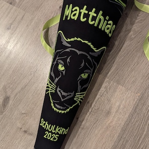 May include: A black and green school cone with a black panther face and green eyes. The cone has the name "Matthias" and the text "Schulkind 2025" embroidered on it.
