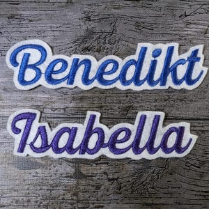 Peut inclure: Deux patchs en tissu blanc avec des lettres brodées bleues et violettes. Le patch du haut indique "Benedikt" et le patch du bas indique "Isabella".