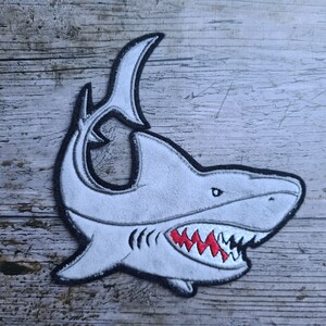 May include: A white embroidered shark patch with a black outline and red teeth. The shark is facing right and has a large open mouth.