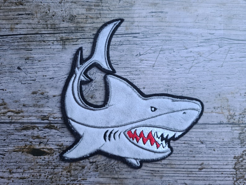 May include: A white embroidered shark patch with a black outline and red teeth. The shark is facing right and has a large open mouth.