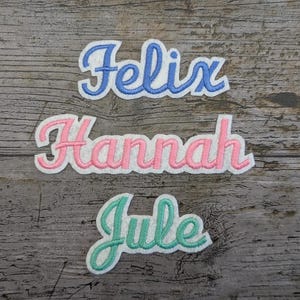 May include: Three embroidered names, Felix, Hannah, and Jule, in different colors on a wood background.