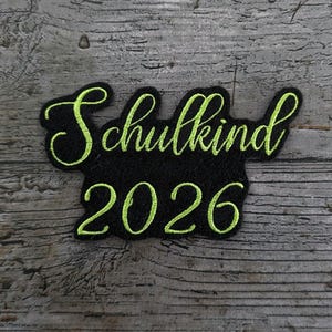 Pode incluir: Um patch bordado preto com as palavras "Schulkind 2026" em uma fonte de script verde neon. O patch está em uma superfície de madeira cinza.