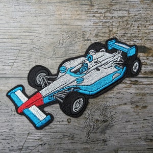 Puede incluir: Un parche bordado de un coche de carreras, con colores azul, blanco y rojo. El parche se muestra sobre una superficie de madera.