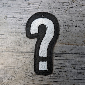 May include: Black and white embroidered question mark patch. The question mark is white, with a black border. The patch has a small hole at the top for attachment. The background is a weathered, gray wooden surface.