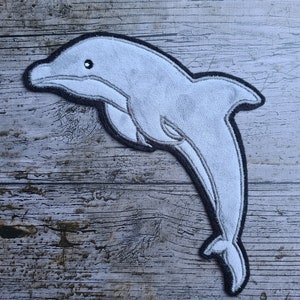May include: A gray felt dolphin applique with black stitching. The dolphin is leaping out of the water with its mouth open.