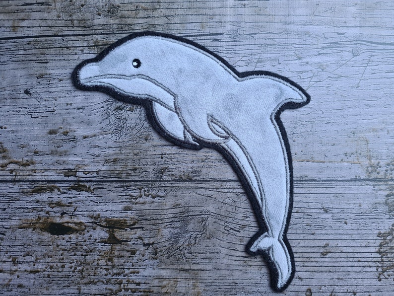 May include: A gray felt dolphin applique with black stitching. The dolphin is leaping out of the water with its mouth open.