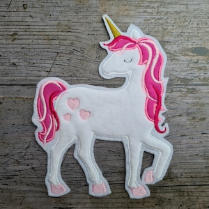 Może przedstawiać: Biała aplikacja z jednorożcem o różowej grzywie i ogonie oraz złotym rogiem. Jednorożec ma trzy różowe serca na boku.