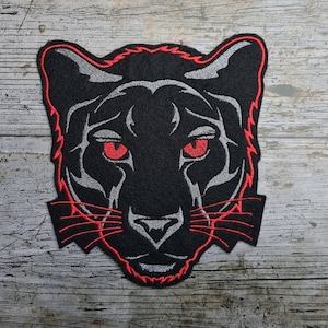 Pode incluir: Patch preto bordado com a cabeça de uma pantera com olhos vermelhos e contorno vermelho. O patch está sobre um fundo de madeira.