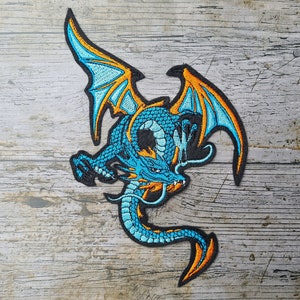 Könnte beinhalten: Ein gestickter Drachen-Patch in Blau und Orange, mit ausgebreiteten Flügeln. Der Drache befindet sich in einer Flugpose mit dem Schwanz um seinen Körper gewickelt.