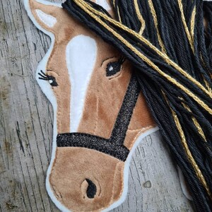 May include: A brown horse head with a white star on its forehead, black eyes, and a black halter with glitter. The horse has a long black mane with gold accents.