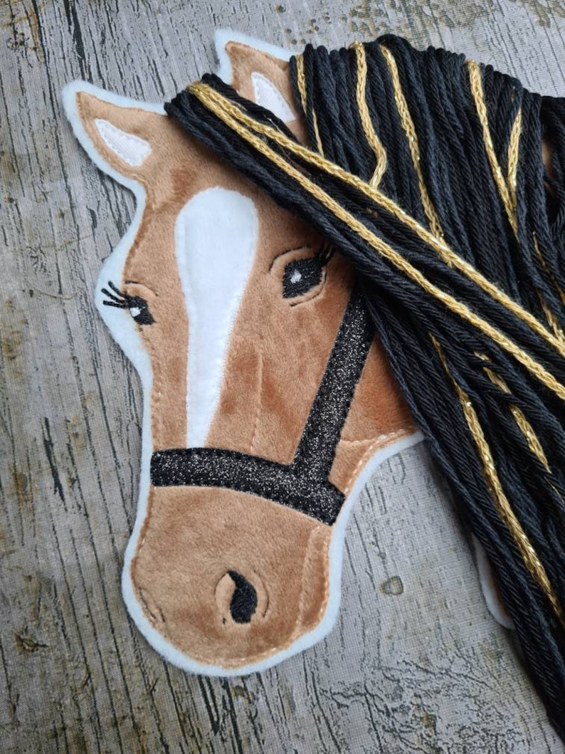 May include: A brown horse head with a white star on its forehead, black eyes, and a black halter with glitter. The horse has a long black mane with gold accents.