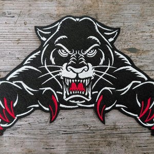 May include: Black panther patch with white and red detailing. The patch depicts an aggressive panther design with sharp claws and teeth. The panther is embroidered with white outlines and red claws, set against a black background.