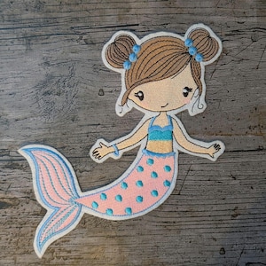 May include: Embroidered patch depicting a cartoon mermaid with brown hair in two buns, decorated with blue details. The mermaid wears a blue and pink top and a pink tail with blue polka dots and trim. Ideal for clothing or craft projects.