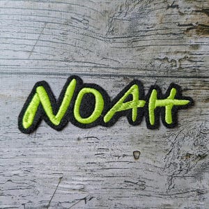 Könnte beinhalten: Ein schwarzer, gestickter Aufnäher mit dem Namen "NOAH" in leuchtendem Grün, umrandet von Schwarz. Der Aufnäher befindet sich auf einer strukturierten, grauen Holzoberfläche. Die Schrift hat einen handschriftlichen Stil.