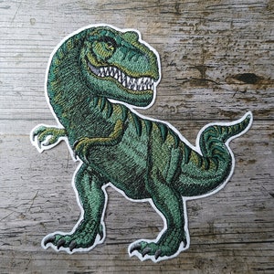 May include: Green embroidered dinosaur patch with white stitching. The dinosaur is standing on all fours with its mouth open and teeth showing.