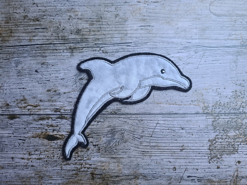 May include: A gray felt dolphin applique with black stitching, on a light gray wood background.
