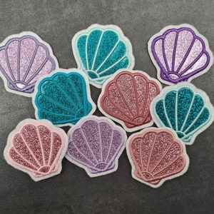 May include: Assortment of eight sparkly seashell patches in various colors, including pink, purple, teal, and blue. The patches are made of fabric and have a glittery finish.
