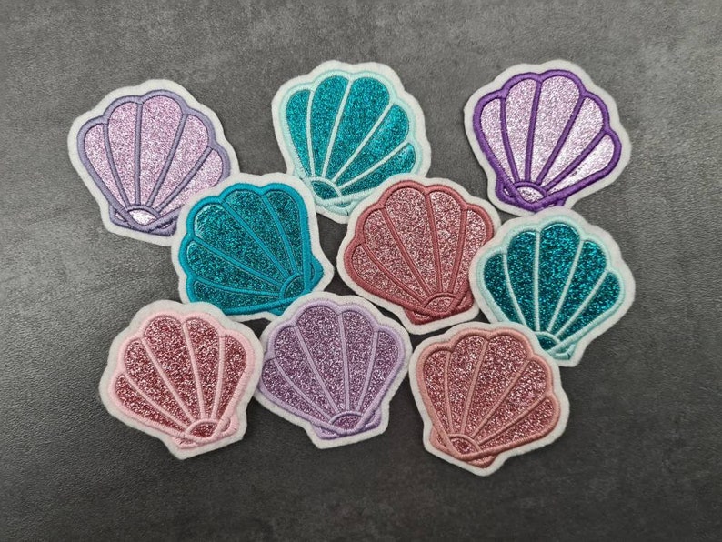 May include: Assortment of eight sparkly seashell patches in various colors, including pink, purple, teal, and blue. The patches are made of fabric and have a glittery finish.