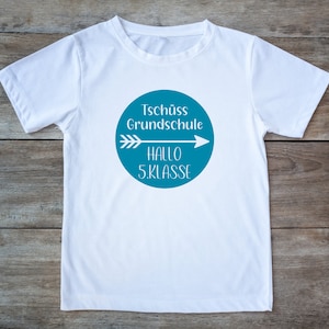 Puede incluir: Camiseta blanca con un gráfico circular azul. El gráfico presenta una flecha blanca apuntando hacia la derecha con el texto "Tschüss Grundschule" sobre la flecha y "Hallo 5. Klasse" debajo de la flecha.